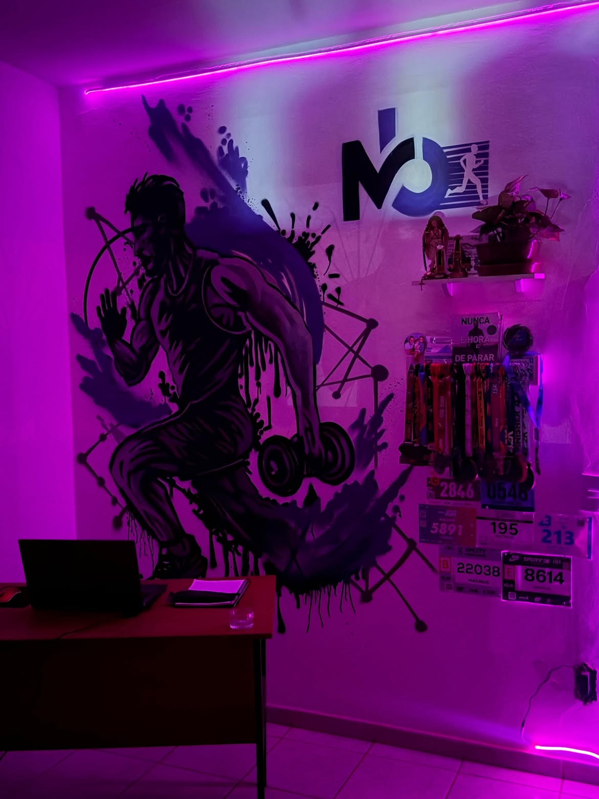 O espaço da MB Performance: mural do corredor com halter, logo MB na parede, medalhas de prova e números de peito sob luz de LED roxa